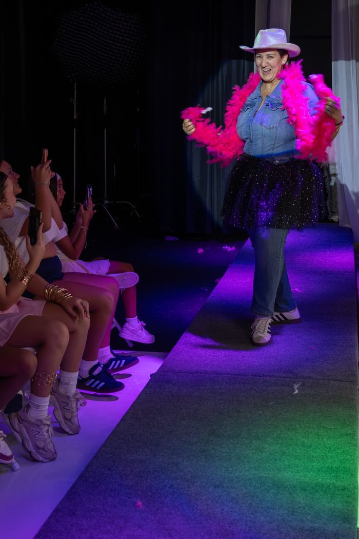 A parent taking a turn on the runway in a pink boa and cowboy hat while girls cheer