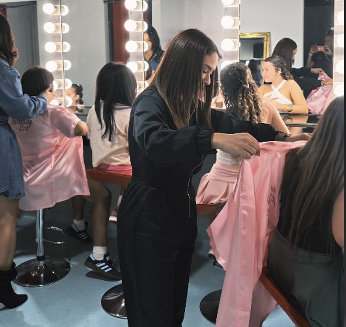 Girls in pink robes getting settled at the vanity mirrors as staff help with their robes